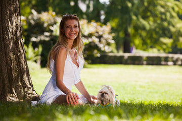 A joyful woman in a summer dress sitting in a lush green park with her adorable dog, soaking up the sunshine.