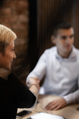 In a warmly lit office with a rustic brick background, a businesswoman shakes hands with a blurred male colleague, signaling a successful meeting or agreement.