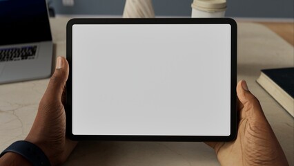 A person holding a tablet with a blank screen, sitting at a desk. Mockup for apps like note-taking,...