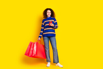 Full size photo of lovely girl dressed sweater holding shopping bags smartphone read notification isolated on yellow color background