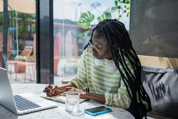 Young professional is working remotely at a cafe, taking notes in a notebook while using a laptop....