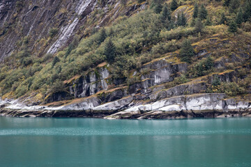 Glacier carved cliff landscape with strong diagonal striations, covered by wild plant life. Variation of color in stone lightness contrasts starkly with a strong aqua water plane.