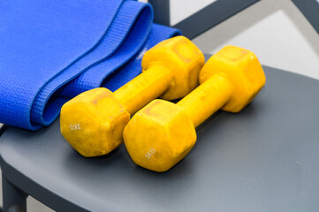 Bright Yellow Dumbbells Resting on a Gray Chair Next to a Rolled Blue Yoga Mat in a Modern Fitness Space