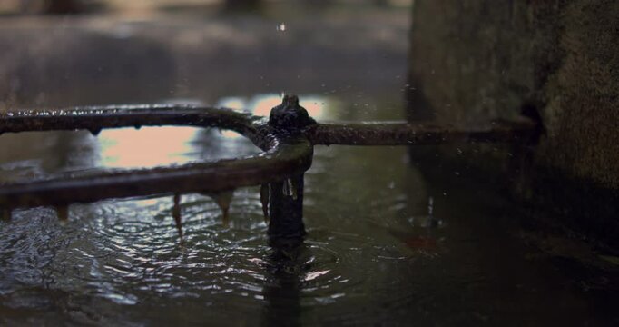 Water dripping from a rusted metal pipe into a fountain, slow motion at 800 fps, close-up of water flow and ripples, peaceful and serene moment in a public fountain