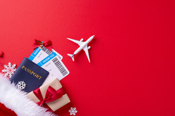 Top view of a holiday travel setup featuring a passport, boarding passes, gift, and airplane over a red background, symbolizing festive travel plans