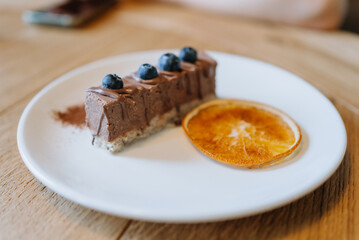 Indulge in a decadent chocolate cake with blueberries and orange on a white plate in a cozy cafe. Rich, creamy, sweet, and tangy, perfect for a snack. Wooden background adds warmth