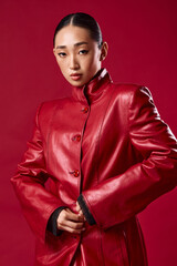 Fashion asian brunette woman in red leather jacket posing with hands on hips and smiling against red background