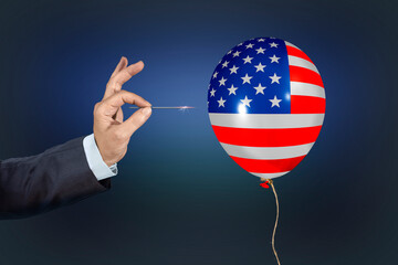 Man holding needle directed to air balloon with the US flag. Concept of risk, loss, catastrophic failure. Hand popping or piercing a balloon with a dart, ready to burst the bubble