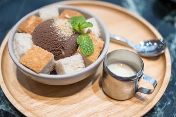 A delectable chocolate ice-cream served with sliced bread and a shot of sweetened condensed milk on a wooden plate is placed on a wooden table at a cafe. Dessert food menu photo, selective focus.