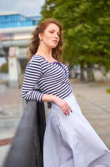 A woman in a striped shirt and white skirt stands on a pier against the backdrop of tall buildings.