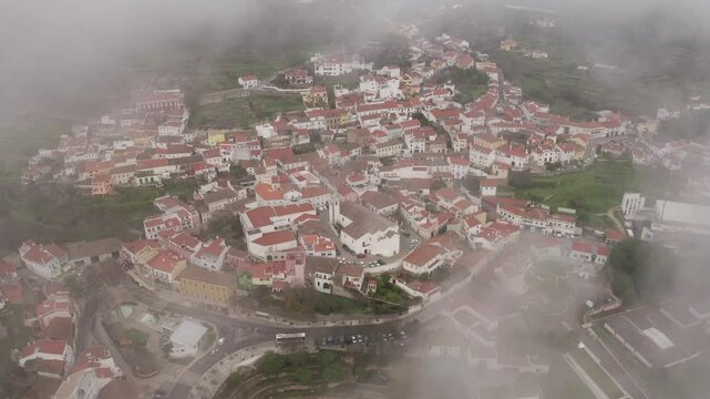 Drone shot of the fog-covered town of Monchique, capturing colorful rooftops and historic buildings. Monchique, Portugal.