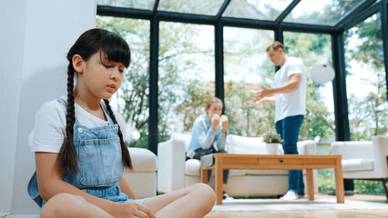 Stressed and unhappy young girl huddle in corner, cover her ears blocking sound of her parent arguing in background. Domestic violence at home and traumatic childhood develop to depression. Synchronos