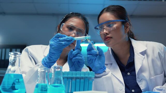 Two female scientists in lab coats and safety glasses are carefully examining a substance in a test tube