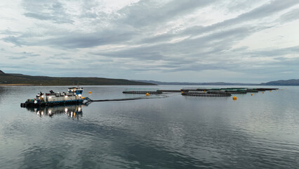 Obraz premium A workboat is involved in aquaculture, floating on calm waters surrounded by mountains and under a cloudy sky.