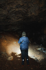 Obraz premium A solitary figure stands quietly in a dimly lit cave, holding a flashlight that casts light on the rocky walls and surrounding shadows, creating an intriguing and captivating air of mystery