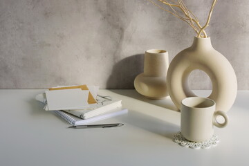 Home office workspace with vases, blank card mockup, envelope, notebooks on white table, gray wall template, Copy space.
