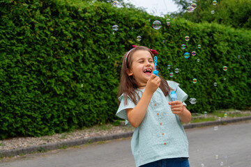 Fototapeta premium Little girl playing with soap bubbles against the background of a green hedge.