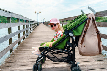 A Caucasian toddle child r girl is sitting in a stroller with sunglasses on an ecotrope on a wooden bridge. Yantarny, Kaliningrad, Russia - 23 June 2024
