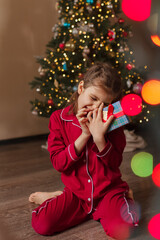 Happy girl of 7 years old in red pajamas sits on living room floor with gifts box next to Christmas tree