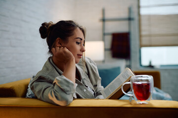 Relaxed woman drinking tea while reading book on sofa.