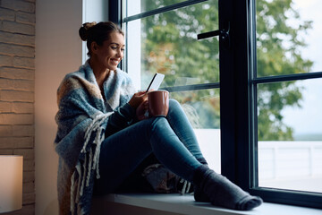 Happy woman using cell phone while drinking tea by window at home.