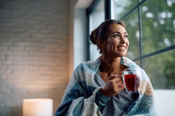 Smiling woman drinking tea while relaxing by window and looking through it.