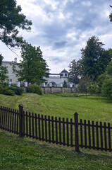 Castle garden fenced with a low wooden fence. Castle building in the background. Trees, grass and sky with clouds. Castle with garden, castle park, travel, monuments.