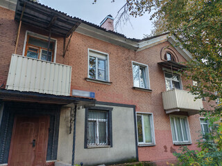 A modest residential building made of red brick, with balconies and a gated entrance, nestled among trees. The scene reflects the character of urban housing from a past era, offering a glimpse into