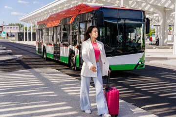 Young girl traveler with a pink suitcase walking along a bus station or airport with a bus in the background.