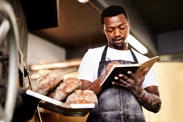 Black man, pastry baker and writing with bread for ingredients, new recipe or texture at factory. Young African, male person or chef with notes for added flavor, mixture or wheat production at bakery