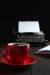 A red cup with coffee in close-up. An old vintage typewriter and an empty piece of paper. A notepad for writing and a red pen. The concept of a business breakfast. Morning of the writer.