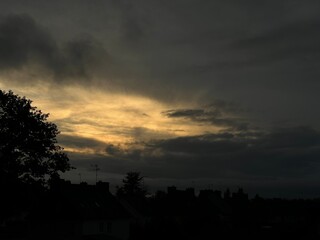 A moody, overcast sky at dusk with dark clouds partially illuminated by a soft golden glow from the setting sun. Silhouettes of rooftops and trees create a dramatic contrast against the fading light.