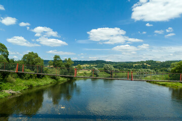 A scenic view of a vibrant blue sky with fluffy white clouds over a serene river, showcasing a picturesque landscape with lush greenery and a red suspension bridge.