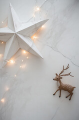 Fototapeta premium A large white wooden star with lights wrapped around it sits on an all-white platform, accompanied by a deer figurine.Minimal creative Christmas holiday concept.Copy space,flat lay