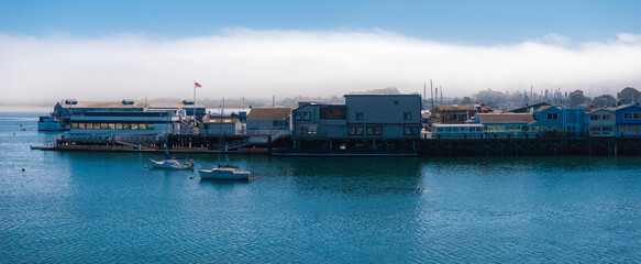 Fototapeta premium Colorful buildings on stilts line the pier in Monterey, California. Small boats are anchored in calm waters, with fog partially obscuring the horizon.