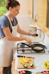 A woman stands at a kitchen stove, wearing an apron and cooking a dish. Fresh vegetables like tomatoes and avocados are neatly arranged on a cutting board nearby, creating a warm atmosphere.
