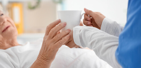 Senior woman taking cup of tea from nurse in bedroom