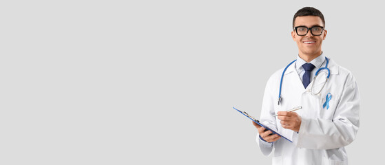 Male urologist with blue ribbon writing in clipboard on light background. Prostate cancer awareness...
