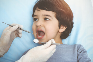 Cute arab boy sitting at dental chair with open mouth during oral checking up with doctor. Visiting sunny dentist office