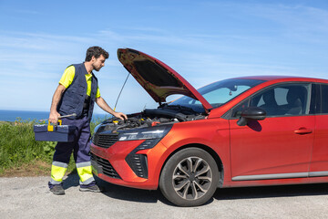 Young mechanic approaching broken vehicle on the road with reflective clothing, toolbox and cross...