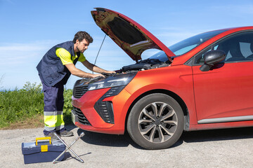 Young mechanic approaching broken vehicle on the road with reflective clothing, toolbox and cross...