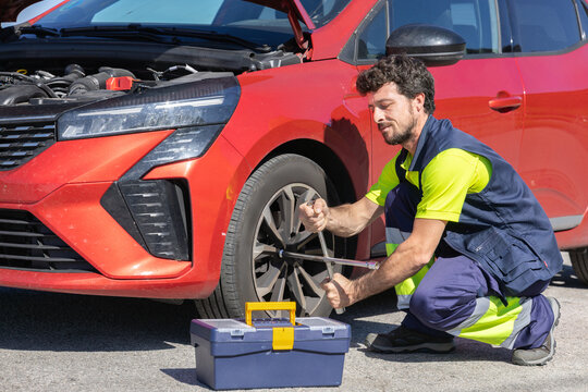 Young professional mechanic changing flat tire broken vehicle on the road with reflective clothing, toolbox and cross wrench, broken engine, roadside assistance, insurance tow truck