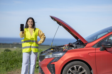 Pretty young woman poses showing phone screen, calling tow truck or insurance mechanic for roadside...