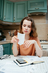 woman pays bills online, sits in kitchen holds receipt in hands with calculator. Online banking at...