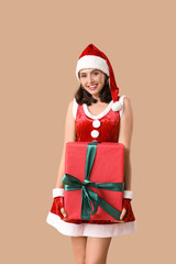 Happy young woman in Santa Claus costume with Christmas gift box on brown background