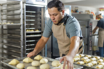 A baker skillfully arranges raw pastry dough on trays, preparing for the baking process in a commercial kitchen setup filled with stainless steel equipment.