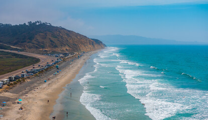 Aerial view of San Diego's coastline with sandy beach, Pacific Ocean, winding road, parked vehicles, rugged cliffs, and people enjoying the sun.