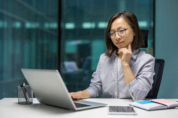 Asian businesswoman deep in thought, working on laptop at office desk. Glasses highlight focus and determination. Laptop, phone, notebook display modern business environment.