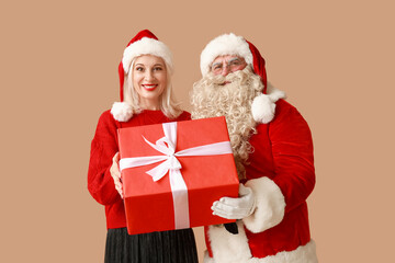 Santa Claus and beautiful mature woman in Christmas hats with gift box on brown background