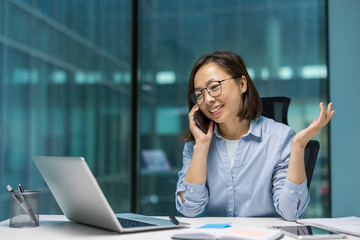 Asian businesswoman on phone call in office. Smiling and confident while using laptop, showcasing modern work environment and communication. Captures professionalism and productivity.